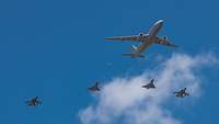 Ein großes Transportflugzeug und vier Kampfjets hinten dran fliegen bei blauem Himmel.