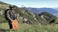 Ein Soldat steht mit orangerotem Signaltuch auf einer Anhöhe im Gebirge. 