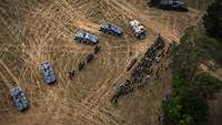 Aus Vogelperspektive sind Militärfahrzeuge, Soldaten und Publikum auf einem braunen Feld zu sehen.