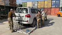 Soldaten sichern ein Auto mithilfe eines Netzes auf einer Palette, im Hintergrund Container