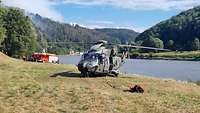 NH-90 mit Löschwasser-Außenlastbehälter steht auf einer Wiese mit See im Hintergrund