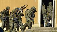 Vier Soldaten mit Gewehren sichern die Hauswand, zwei Soldaten stürmen durch eine Tür.