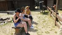 Frauen sitzen mit Sonnen- bzw. Cowboyhüten und kühlen Getränken auf einer Bank.