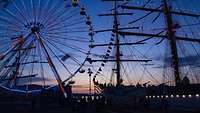 Es sind ein Riesenrad und ein Segelschiff zur Abendstunde zu sehen.