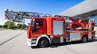 Ein großes rotes Feuerwehrauto mit langer Drehleiter und Rettungskorb steht auf einem großen Platz vor einer Fahrzeughalle