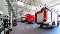 In einer großen Fahrzeughalle stehen drei rote Feuerwehrautos. An den Wänden hängen Feuerwehr-Uniformen an Kleiderhaken.