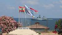 Im Hintergrund fährt die deutsche Korvette, im Vordergrund wehen die Flaggen der Nationen, die sich an UNIFIL beteiligen