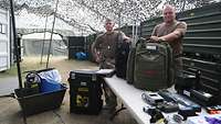 Zwei Soldaten stehen unter einem Tarnnetz an einem Tisch mit Rucksäcken,Transportkoffer und Geräten