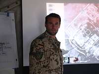 Ein Soldat zeigt mit einem Zeigestock auf eine Beamer-Projektion, die ein Luftbild von einem Gelände zeigt.