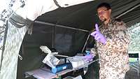 Ein Soldat steht mit erhobenen Zeigefinger in einem Zelt und hält eine Plastiktüte mit Röhrchen mit einer Greifzange
