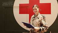 Eine Soldatin steht vor einem Container mit rotem Kreuz auf weißem Grund und hält ein Klemmbrett in den Händen