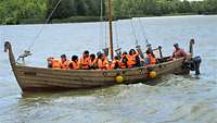 Menschen sitzen mit Rettungswesten in einem nachgebauten Slawenboot und fahren auf einem See.