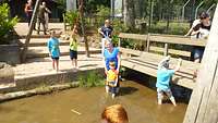 Kinder stehen im und an einem Teich, eine Frau steht ebenso im Wasser.