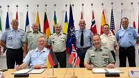 Soldiers of different nations stand in front of national flags. Two soldiers sit at a table