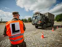 Ein Soldat mit Warweste und Aufschrift „L–Experiment“ steht mit dem Rücken zum Betrachter vor einem LKW auf einer Teststrecke.