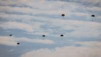 Fünf Fallschirmspringer fliegen am Himmel, links daneben fliegt das Flugzeug