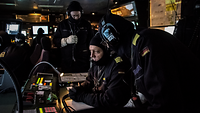 Mehrere Marinesoldaten in blauen Arbeitsanzügen mit blauen Sturmhauben und mit aufgesetzten Kopfhörern.