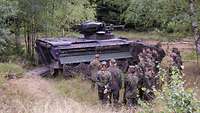 Mehrere Personen in Uniform stehen im Wald vor einen Panzer.