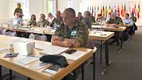 Mehrere Soldatinnen und Soldaten verschiedener Nationen sitzen in einem Raum an Tischen. Im Hintergrund mehrere Nationalfahnen.