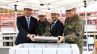 Zwei Soldaten und zwei Männer in Anzügen halten gemeinsam einen Metallzylinder in der Hand. Sie stehen auf einer Baustelle.