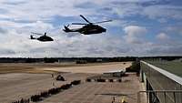 Zwei Hubschrauber fliegen über die Formation zahlreicher Soldaten auf einem Flugplatz.
