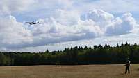 Auf einer Wiese steht ein Soldat mit einer Fernsteuerung. Am Himmel über der Wiese ist eine Drohne zu sehen.