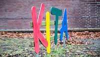 Ein buntes Schild mit der Aufschrift „Kita“ ist vor einer Hauswand zu sehen