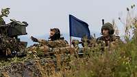 3 Soldaten sitzen auf einem GTK Boxer. In der Mitte weht eine blaue Flagge. Der linke Soldat zeigt mit der Hand nach links.