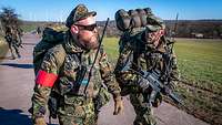 Ein Soldat mit roter Armbinde und ein bewaffneter Soldat laufen auf einer Straße nebeneinander.