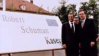 Zwei Zivilisten stehen neben einem weißen Schild mit der Aufschrift: „Robert Schuman Kaserne“.