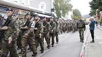 Soldaten marschieren in Formationen in einer Stadt auf der Straße, daneben stehen Zivilisten.