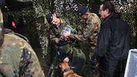 Zwei Soldaten unter einem Tarnnetz ziehen einem Zivilisten einen schweren Schutzanzug an.