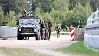 Ein Fahrzeug fährt auf einer Straße neben einem Wald. Dahinter laufen Soldaten, die Deckung suchen.