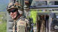 Ein Soldat mit Helm, Gehörschutz, Sonnenbrille und Sicherungsseilen lächelt, im Hintergrund Fahrzeuge
