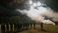 Mehrere Soldaten stehen bei Dunkelheit auf einem Feld. Vor ihnen leuchten grell weiße Lichtquellen und Rauch steigt auf.