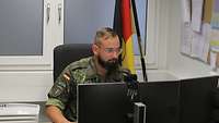 Ein Soldat sitzt an seinem PC-Arbeitsplatz mit zwei Bildschirmen. Im Hintergrund steht eine Bundesdienstflagge.