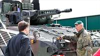 Ein Zivilist in dunklem Anzug spricht vor einem Panzer mit einem Soldaten vor einer Halle.