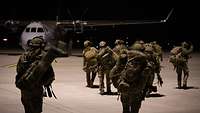 Soldaten verschiedener Nationen gehen mit Einsatzgepäck bei Dunkelheit zu einem wartenden Transportflugzeug.
