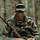 Ein Soldat mit Tarnfarbe und Brille hockt in einem Waldstück: Er hält ein Funkgerät.
