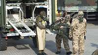 Drei Soldaten in Schutzanzug tragen einen weiteren auf einer Trage. Hinter ihnen ein Bundeswehrsanitätsfahrzeug.