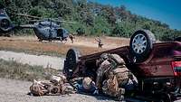 Ein KSK-Soldat kniet neben einem verletzten Soldaten vor einem zerstörten Auto. Dahinter Soldaten und ein Hubschrauber am Boden.