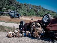 Ein KSK-Soldat kniet neben einem verletzten Soldaten vor einem zerstörten Auto. Dahinter Soldaten und ein Hubschrauber am Boden.