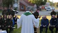 Rückenansicht auf einen Priester, vor ihm sitzen einige Menschen auf Bänken.