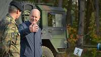 Vor einem grünen Lastwagen im Wald spricht ein Zivilist in einem blauen Mantel einem General.