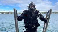 Ein Taucher mit Taucherhelm im schwarzen Neoprenanzug steigt eine Metalltreppe hinab ins Wasser.