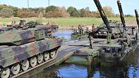 Über eine Schwimmbrücke fahren Panzer von einem Flussufer zum anderen. Dazwischen stehen Soldaten.