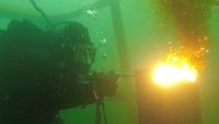 Ein Taucher hält unter Wasser eine thermische Lanze, an deren Ende entsteht eine Stichflamme. Er zielt auf eine Spundwand.