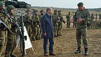 Bundeskanzler Scholz spricht mit einem Soldaten auf einem Feld. Dahinter stehen mehrere Soldaten vor Panzern.