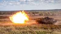 Auf weitem freien Gelände schießt ein Panzer, ein großer gelber Feuerball verlässt die Kanone.