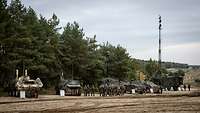 Mehrere militärische Fahrzeuge stehen auf einer großen Sandfläche mit Abstand nebeneinander, davor Infotafeln und Soldaten.